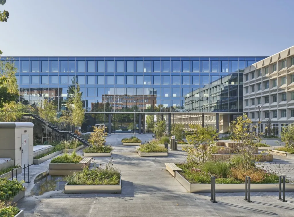 En modern byggnad med glasfasad omgiven av en välskött trädgård med gångvägar och planteringar.