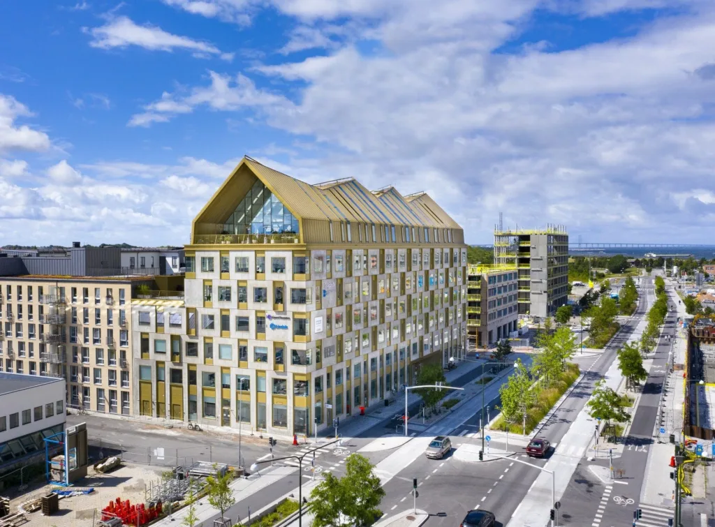Ett modernt flerfamiljshus med trädetaljer och spetsigt tak, omgivet av grönska och trottoarer, under en klarblå himmel.