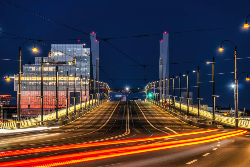 En upplyst bro nattetid med röda och gula ljusstrimmor från trafiken, och höga byggnader i bakgrunden.