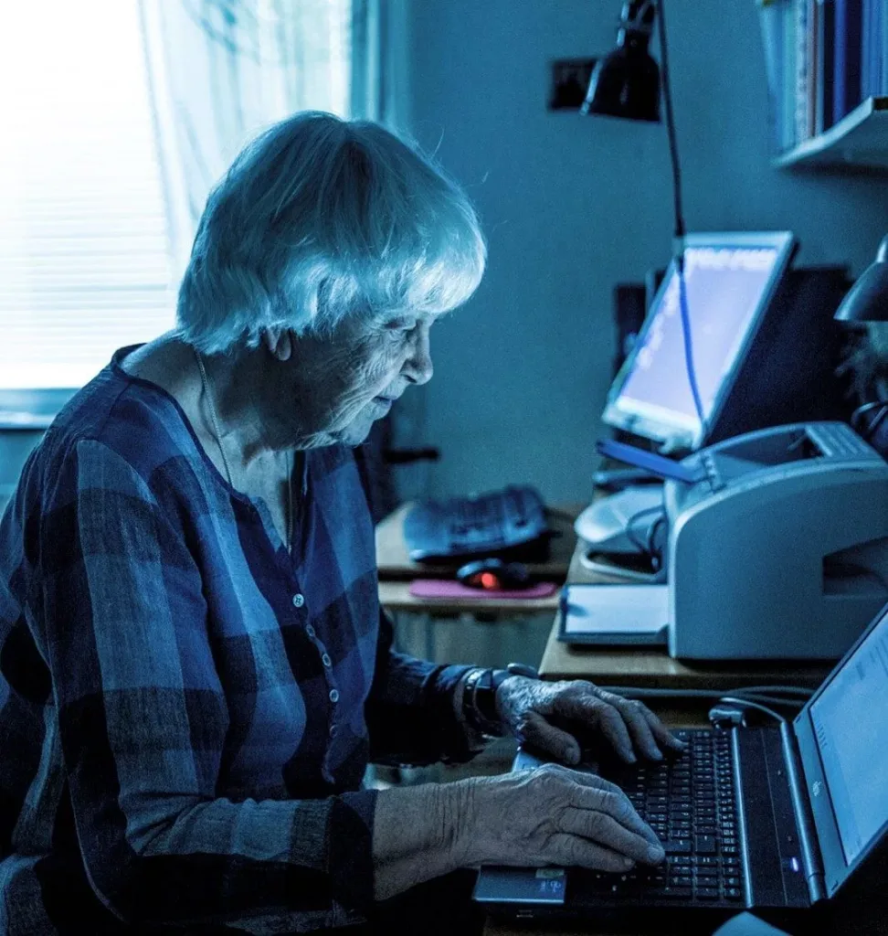 En äldre kvinna sitter vid en bärbar dator i ett rum med blåaktigt ljus, omgiven av skrivare och kontorsmaterial.
