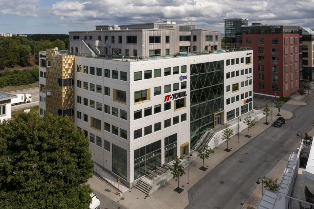En modern byggnad med stora glasfönster och företagsskyltar på fasaden, en bred gata och träd runt omkring.