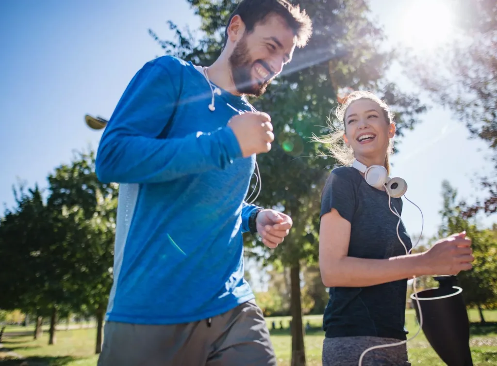Två personer joggar utomhus i solskenet; en man i blå tröja och en kvinna med hörlurar.