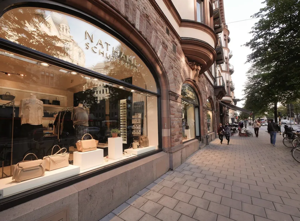 En butik med stora skyltfönster som visar väskor och andra varor, samt en trottoar med gående människor.