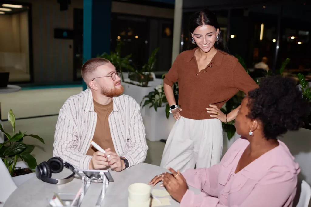 Diverse creative team of two colleagues sitting at cafe table participating in business meeting with supportive female boss checking up on work progress at office center, camera flash