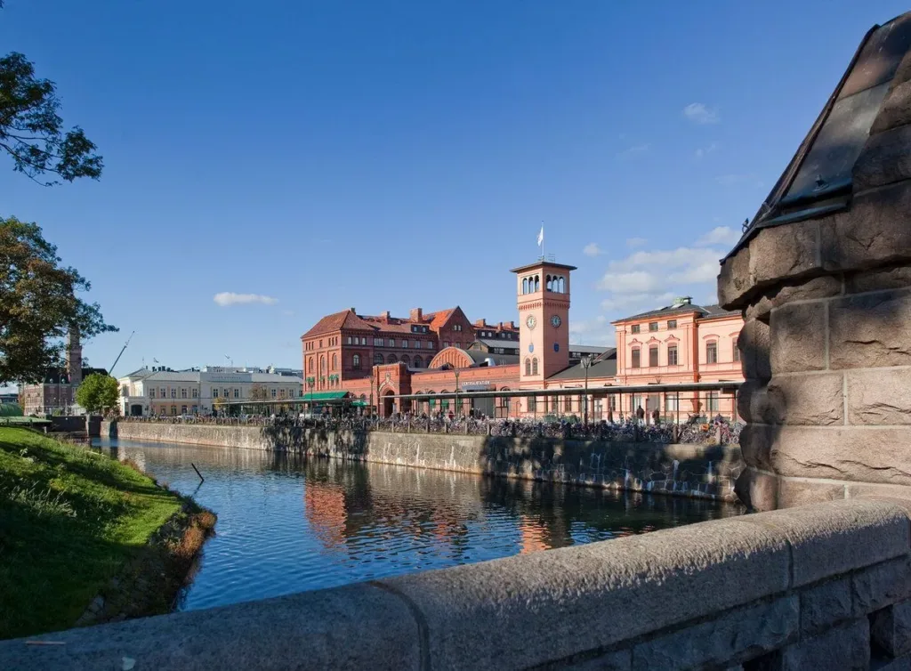 En stadsbild med en flod, röda tegelbyggnader och en klocktorn under en klarblå himmel.