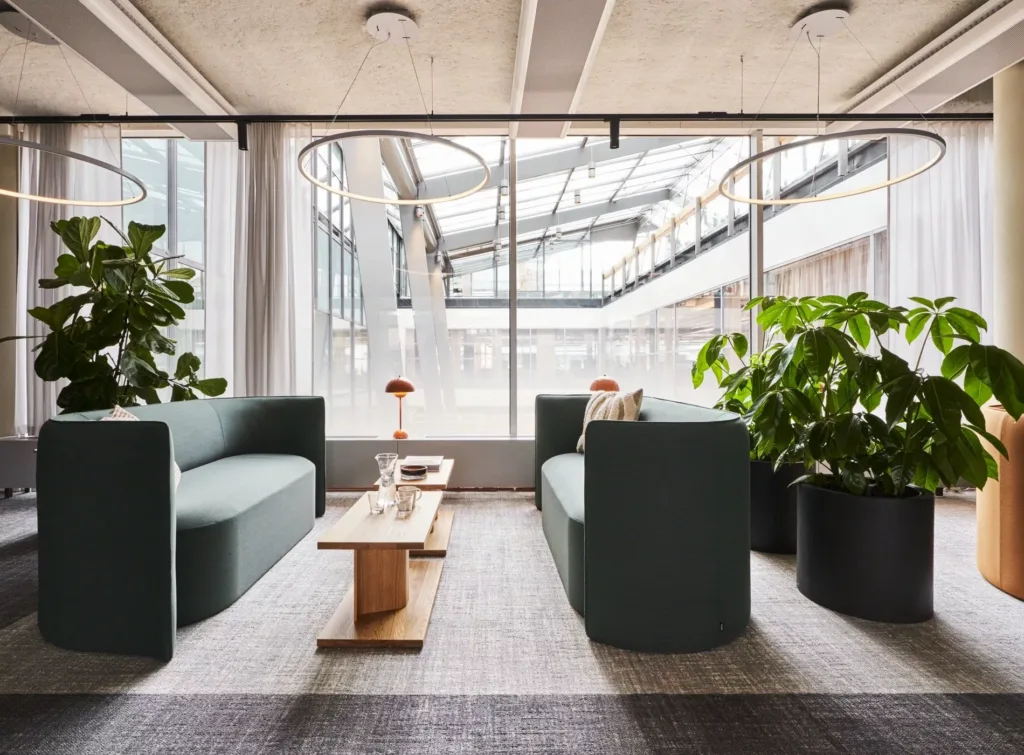 Ett loungeområde med två gröna soffor, ett träbord och stora växter nära fönster med utsikt över en glasfasad.
