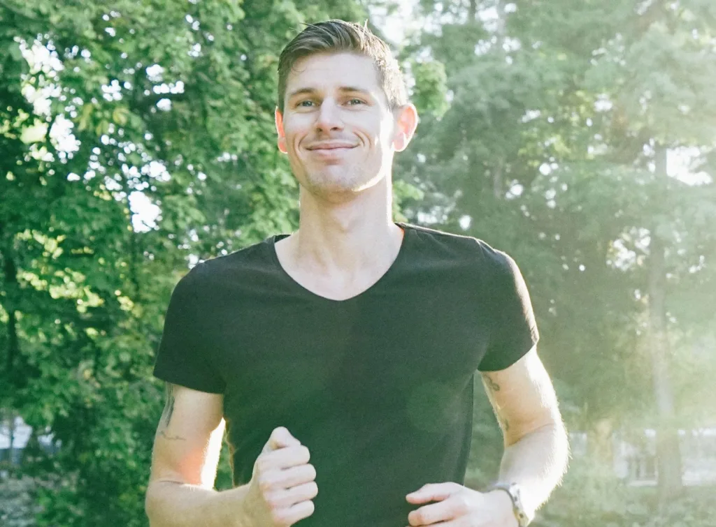 En person klädd i en svart t-shirt joggar i en park med soligt väder och grönska i bakgrunden.