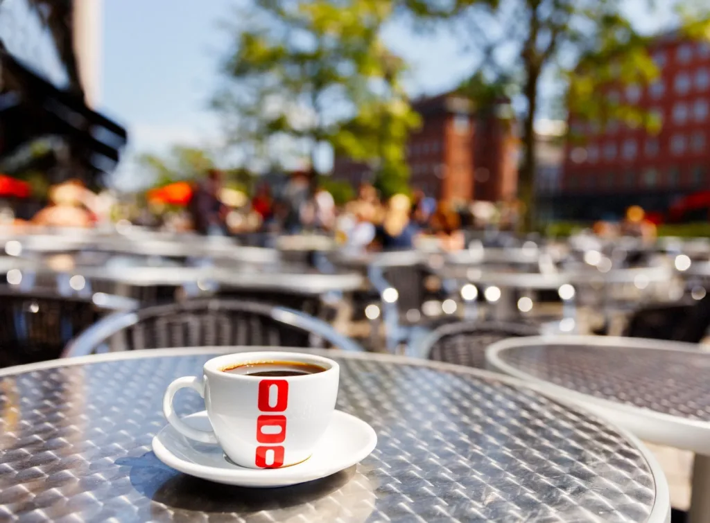 En kopp kaffe med vitt porslin och röd logotyp på ett metallbord utomhus, med suddig bakgrund av uteservering.