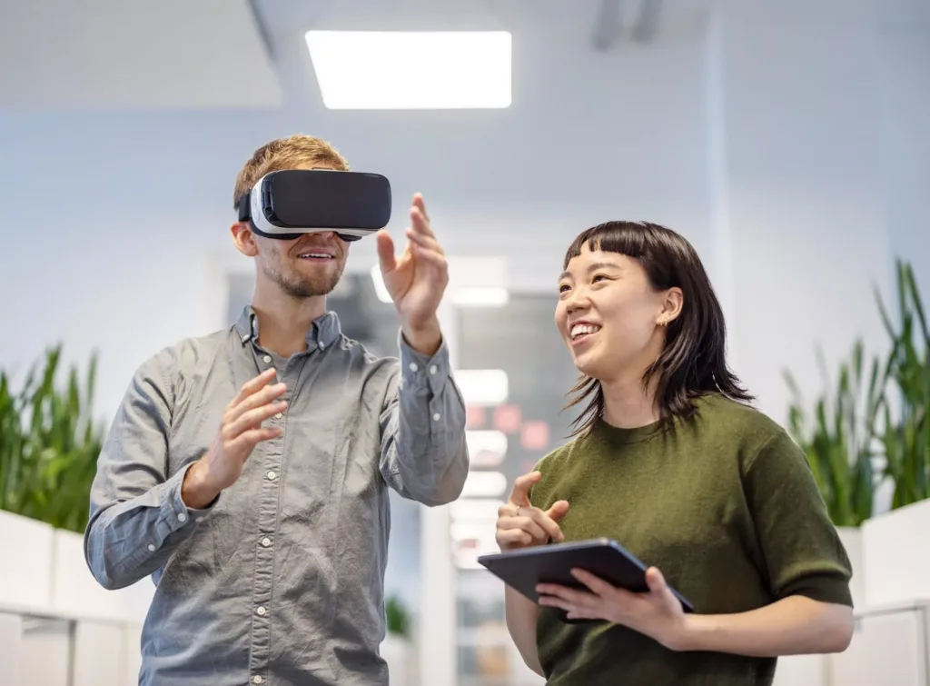 En man med VR-glasögon sträcker ut handen medan en kvinna bredvid honom ler och håller en surfplatta.