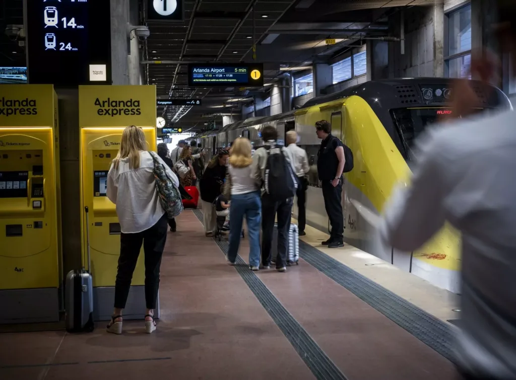 En tågperrong med människor som väntar, en gul automat för biljetter och ett gulsvart tåg.