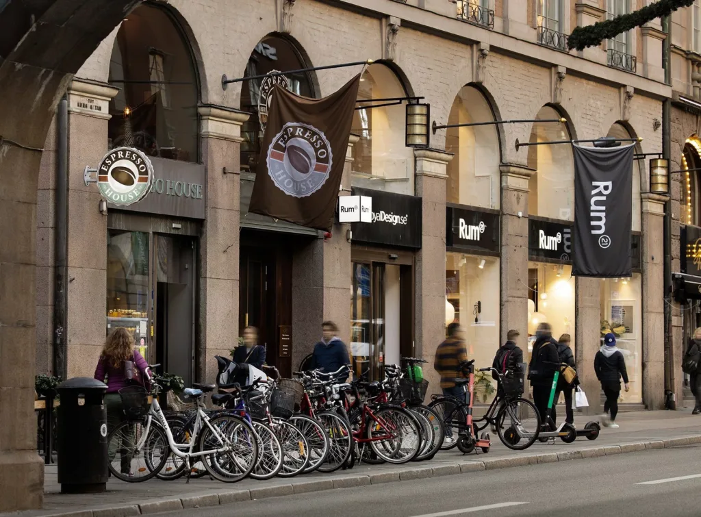 En rad av människor och cyklar framför en byggnad med affärsskyltar och flaggor.
