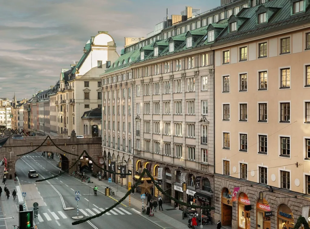 En rad av äldre byggnader med gröna tak och valv längs en stadsgata, bilar och fotgängare synliga.
