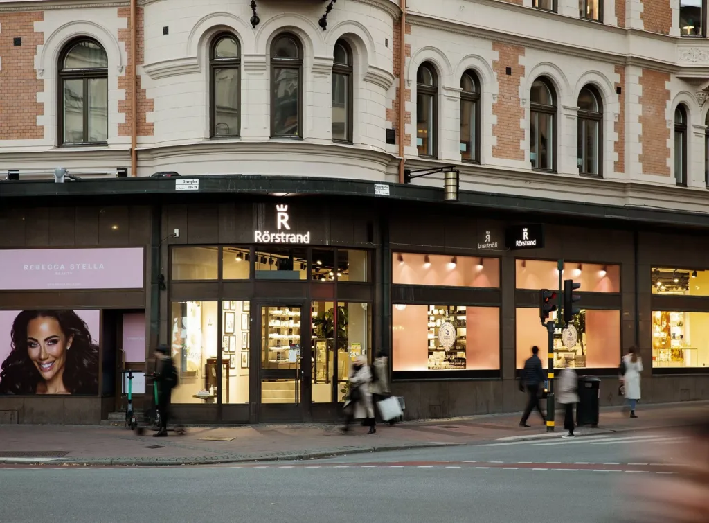 En butik med skyltar som lyser upp i kvällsljuset, människor går förbi på trottoaren.