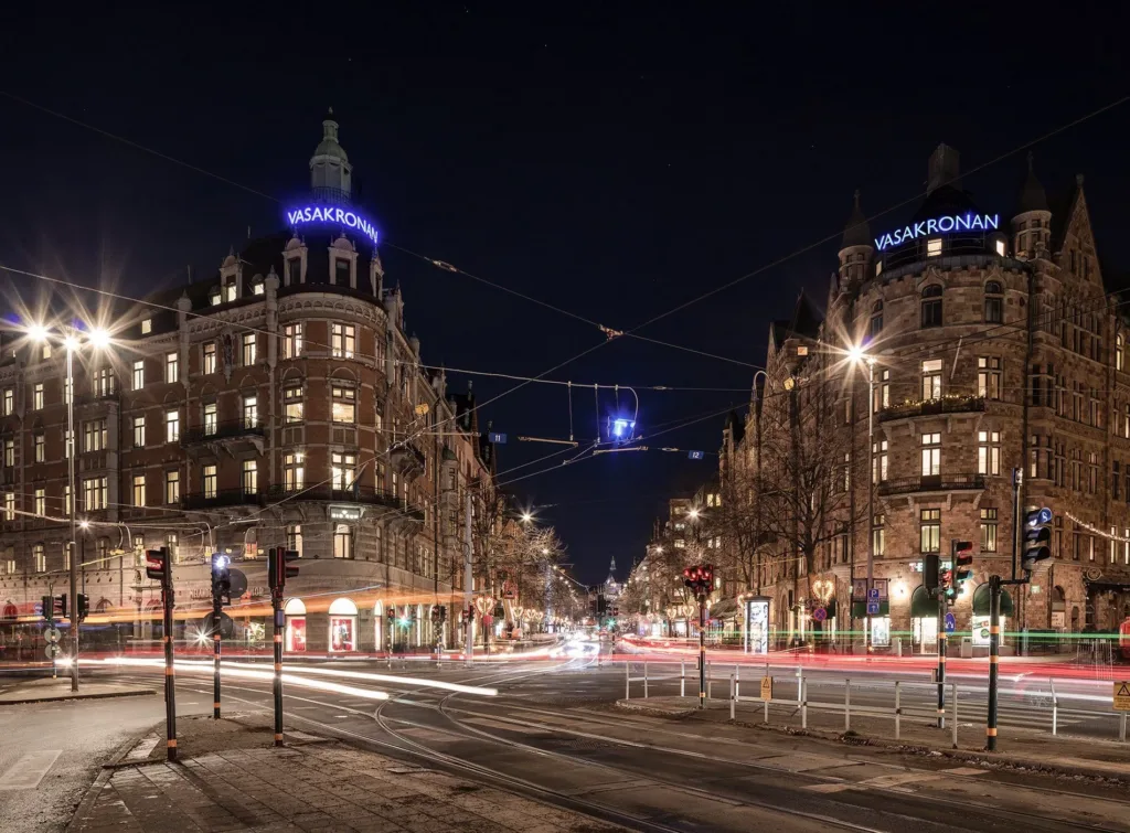 En stadsgata på kvällen med byggnader upplysta och bilars ljusspår synliga.