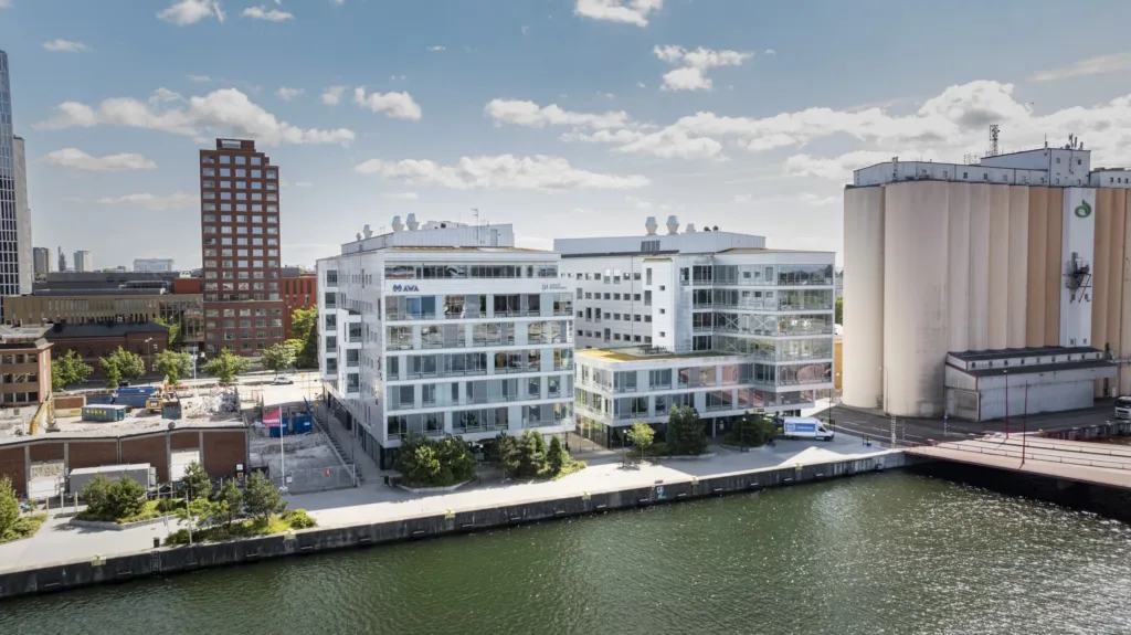 Moderna byggnader vid en kanal, omgivna av träd och blå himmel.