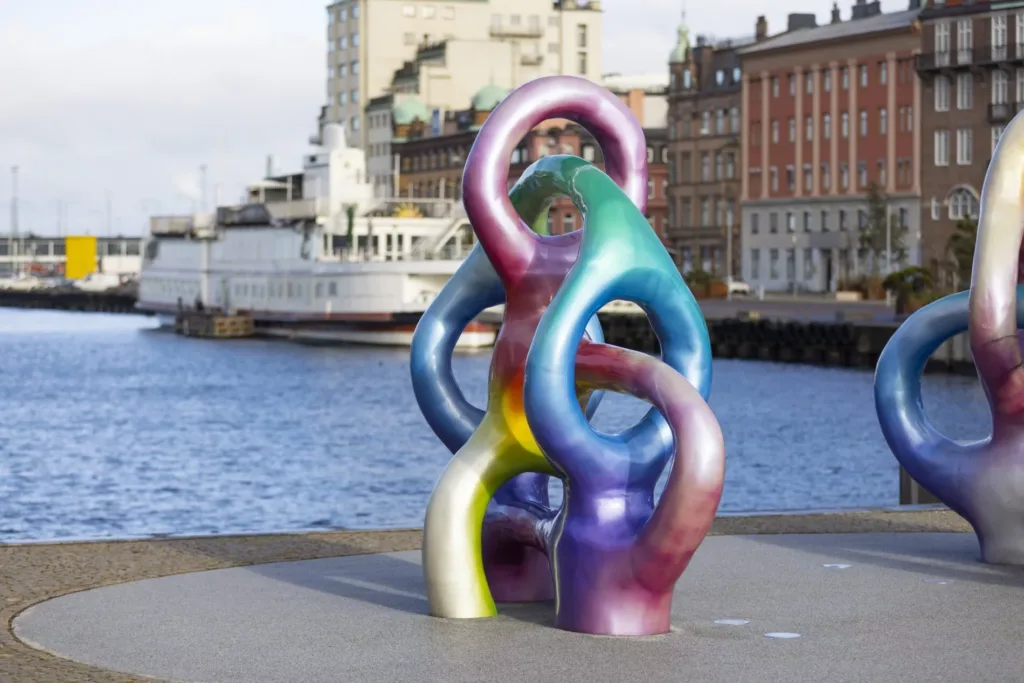 En färgglad skulptur med sammanflätade former vid vattnet i en stad.