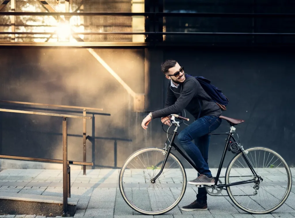 En man med solglasögon cyklar på en stadsgata med solstrålar som lyser bakom honom.