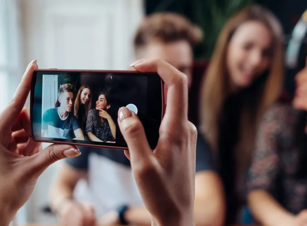 En hand håller en mobiltelefon som tar en selfie på tre personer som ler.