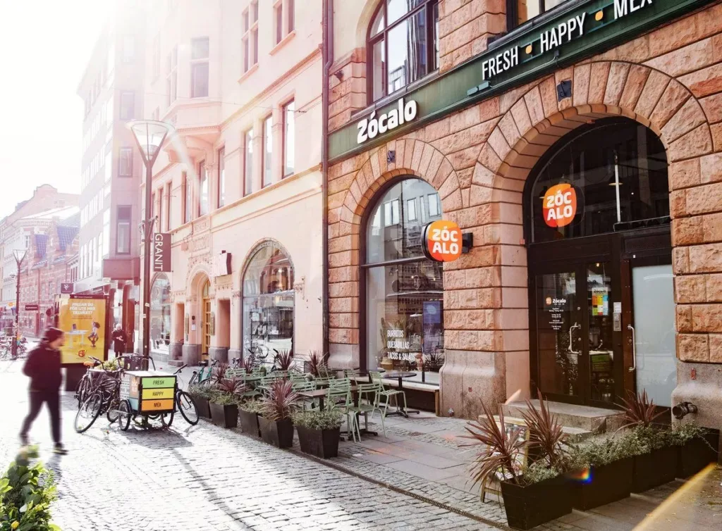 En solbelyst gata med uteservering, bord och stolar framför en byggnad med bågformade fönster.