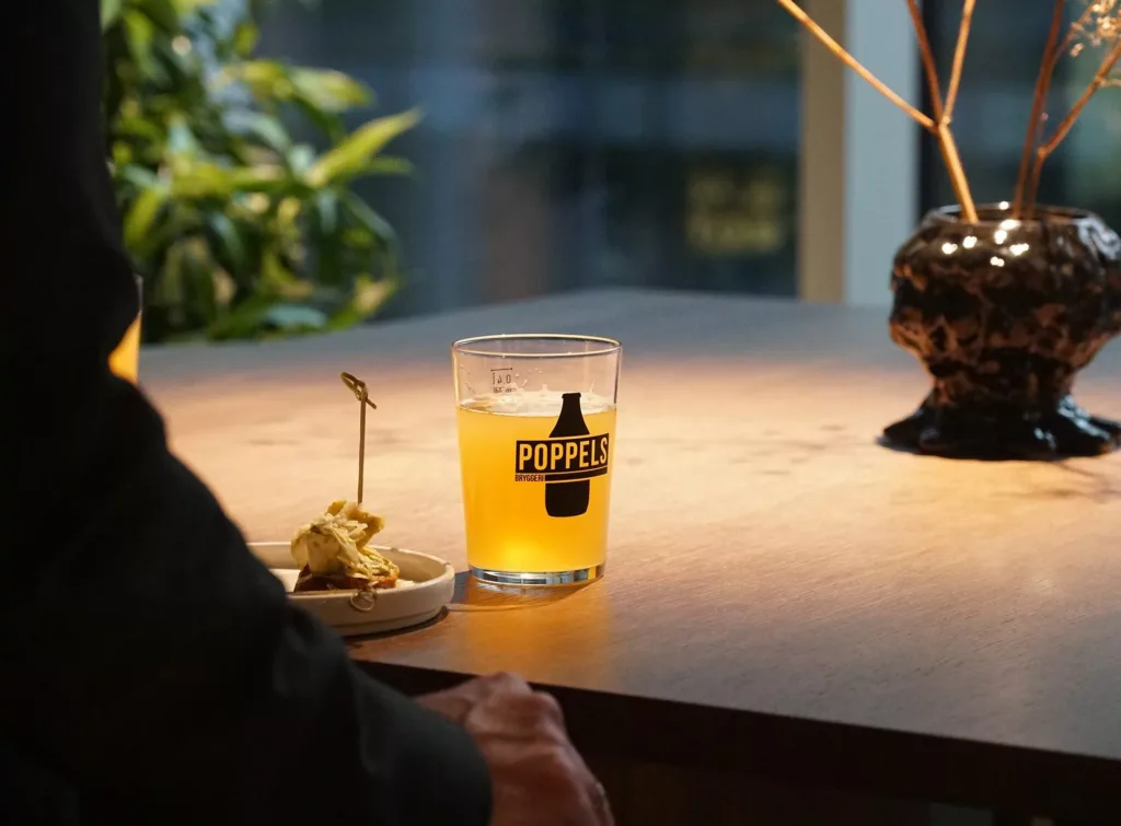 Ett glas öl med texten "KAPPA" på en träbord, bredvid en tallrik med mat.