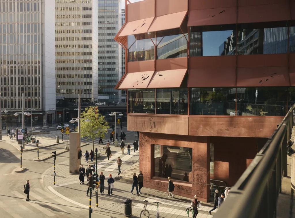 En modern byggnad med orange fasad och glasfönster, omgiven av människor och stadsmiljö.
