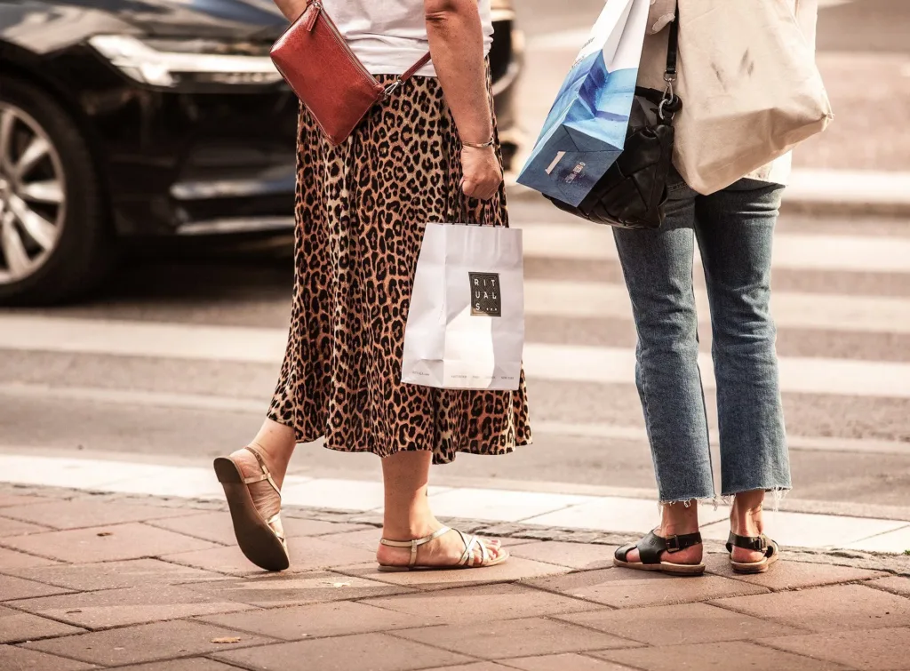 Två personer står på en trottoar vid ett övergångsställe med shoppingpåsar i händerna.