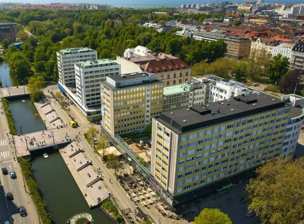 Drönarvy över byggnader, grönområden och en kanal med bryggor i en stadsmiljö.