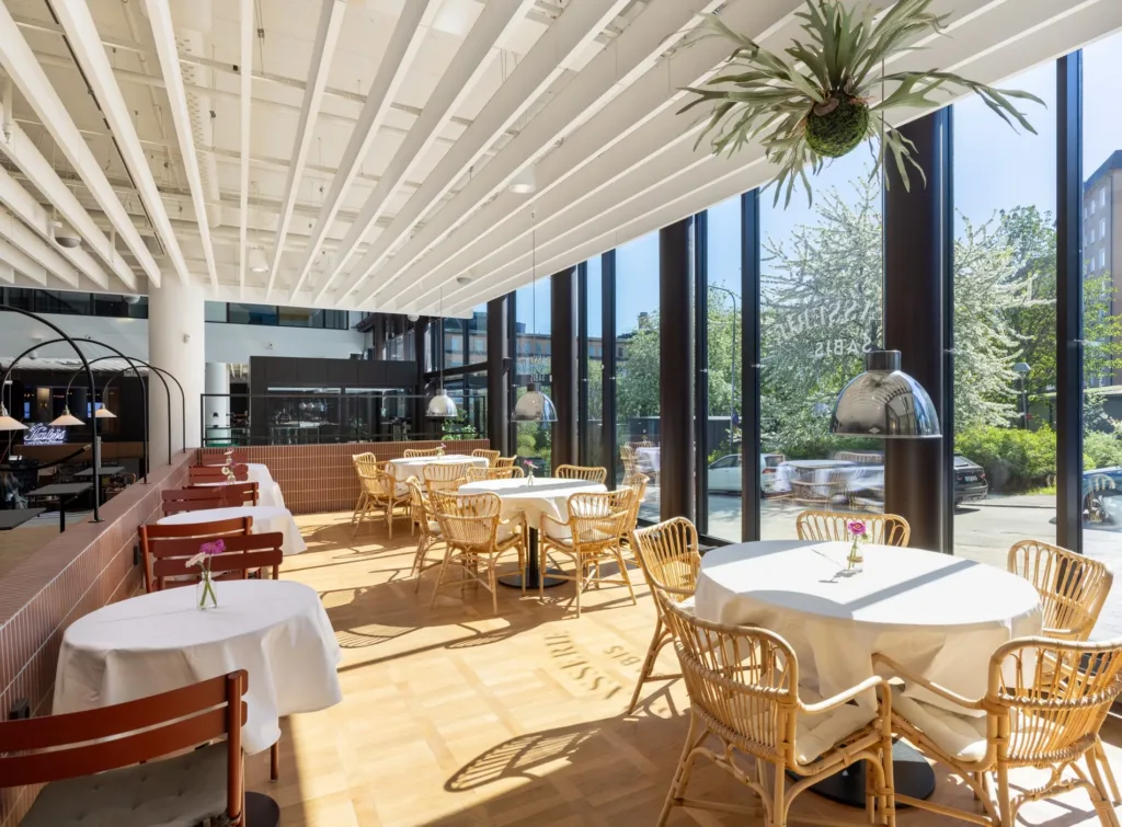 En ljus restaurang med stora fönster, trästolar och bord med vita dukar.