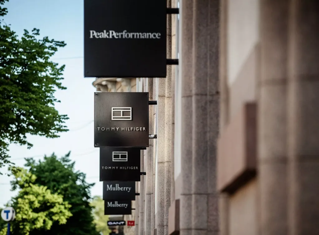Flera svarta butiksskyltar med märkesnamn, inklusive "Peak Performance" och "Mulberry", längs en rad byggnader.
