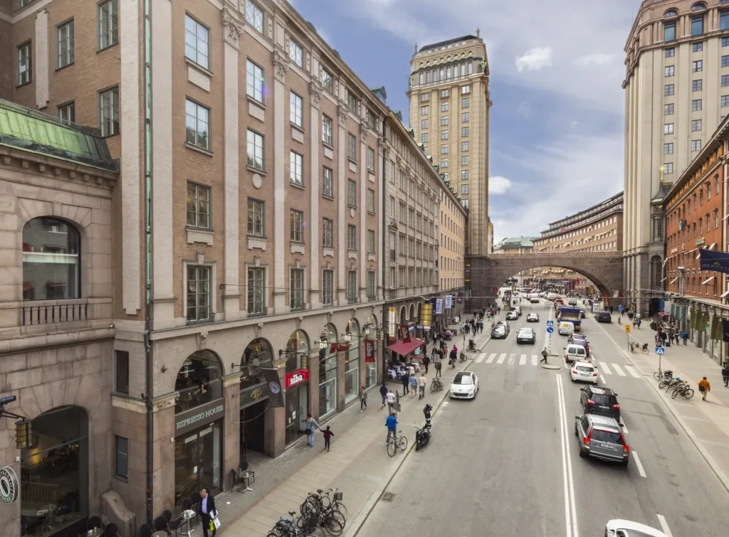 En livlig stadsgata med höga byggnader, fotgängare och bilar under en bro i bakgrunden.