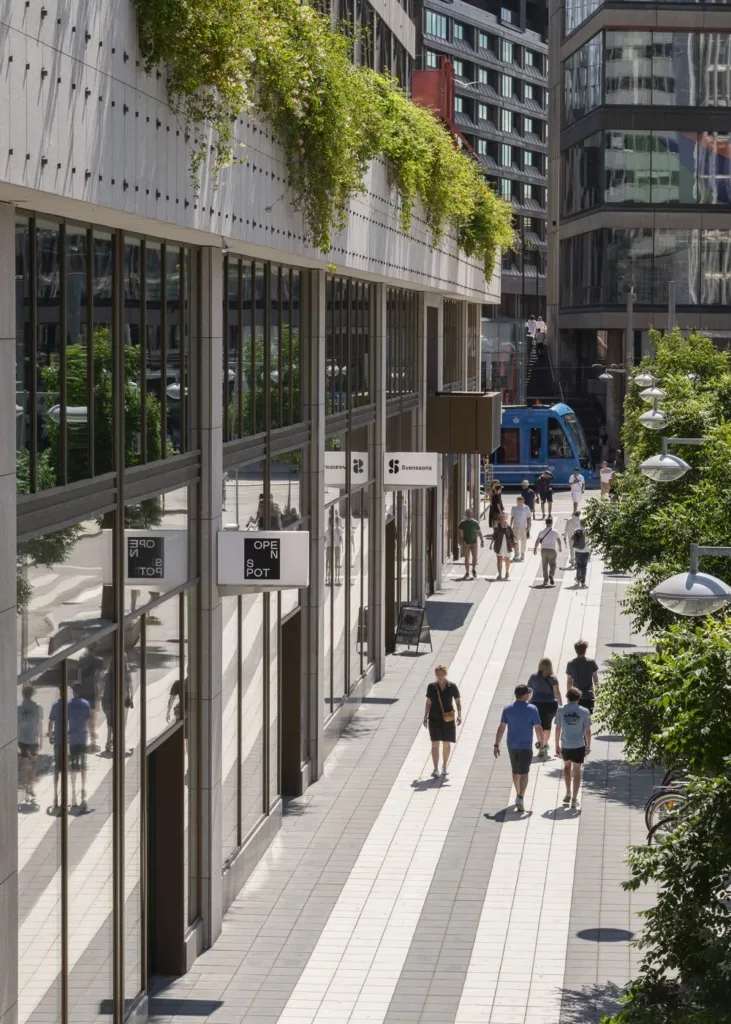 En bred gångväg kantad av byggnader med glasfasader och grönska, flera personer promenerar och en blå spårvagn syns i bakgrunden.
