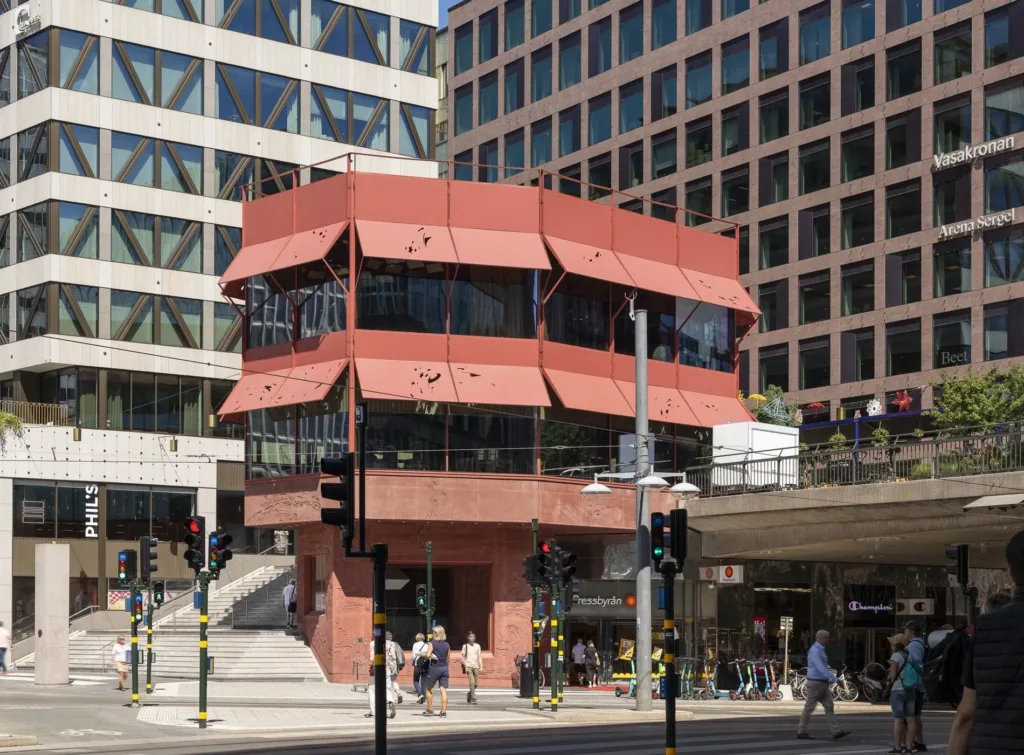 En röd byggnad med geometrisk design omgiven av gångvägar och trafikljus, människor rör sig på gatan.