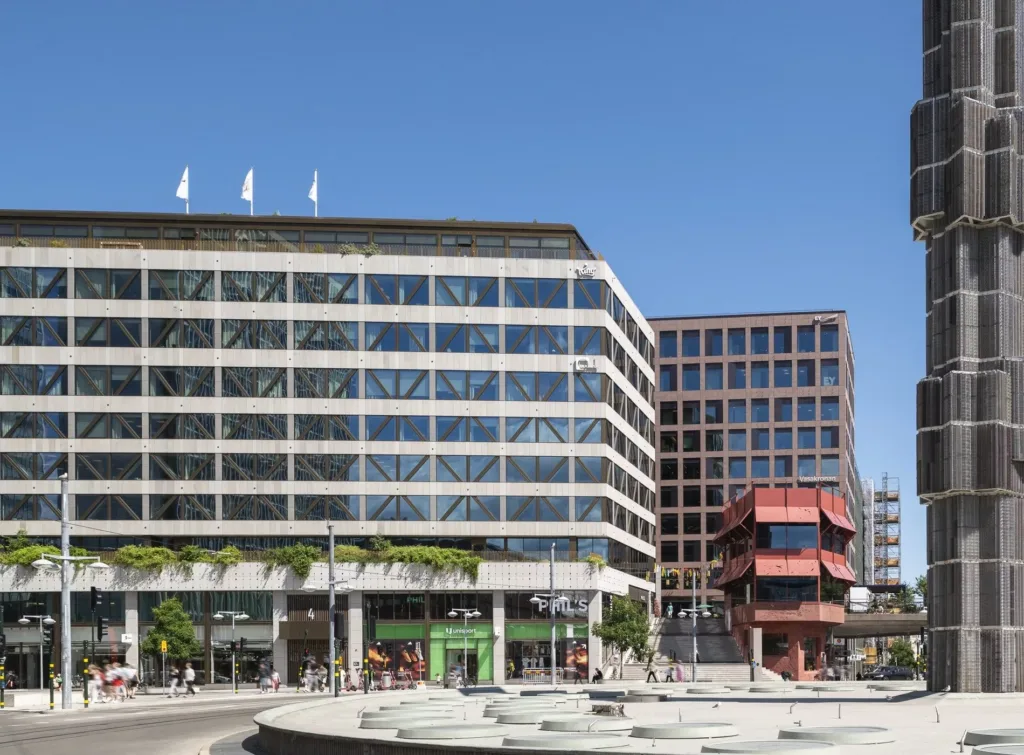En modern byggnad med stora glasfasader och en mindre röd byggnad bredvid, omgiven av en stadsmiljö med blå himmel.