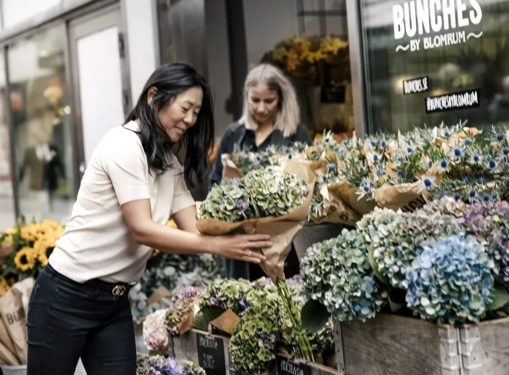 Två kvinnor står vid en blomsterbutik och tittar på färgglada buketter.