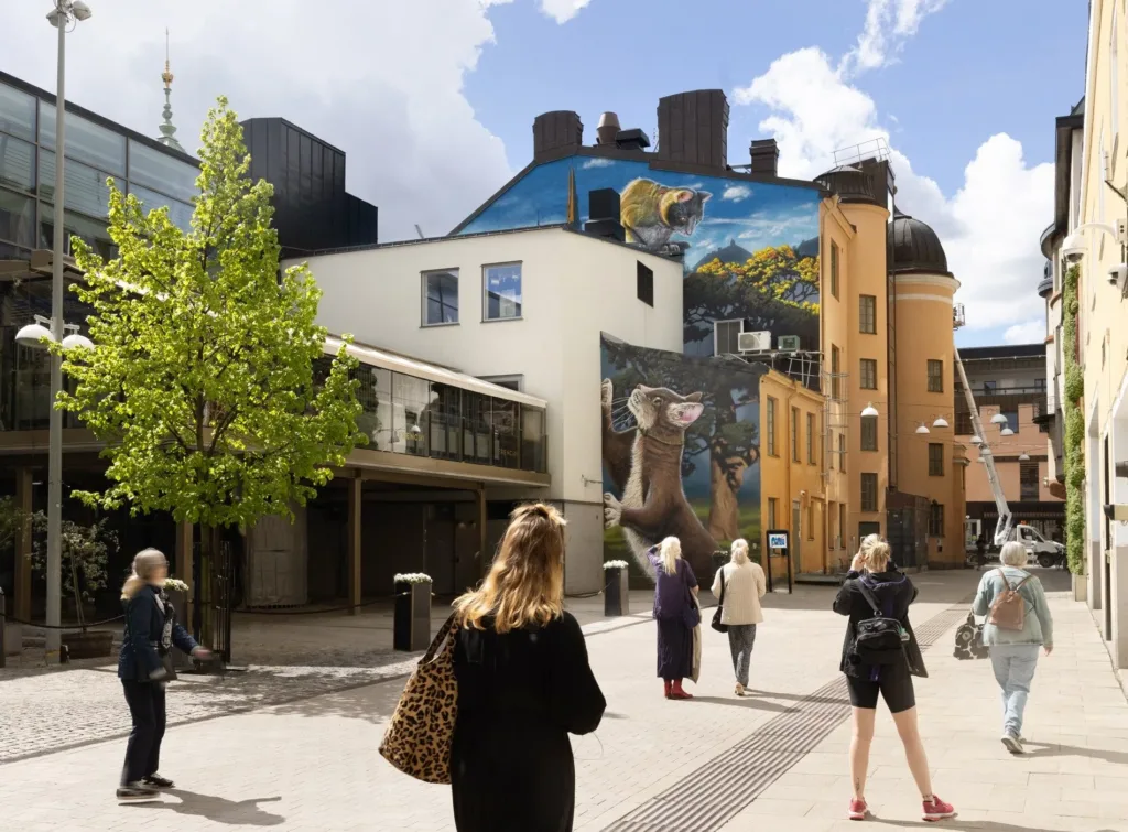 Gågata med personer som promenerar, väggmålning med djurmotiv på en byggnad i bakgrunden.