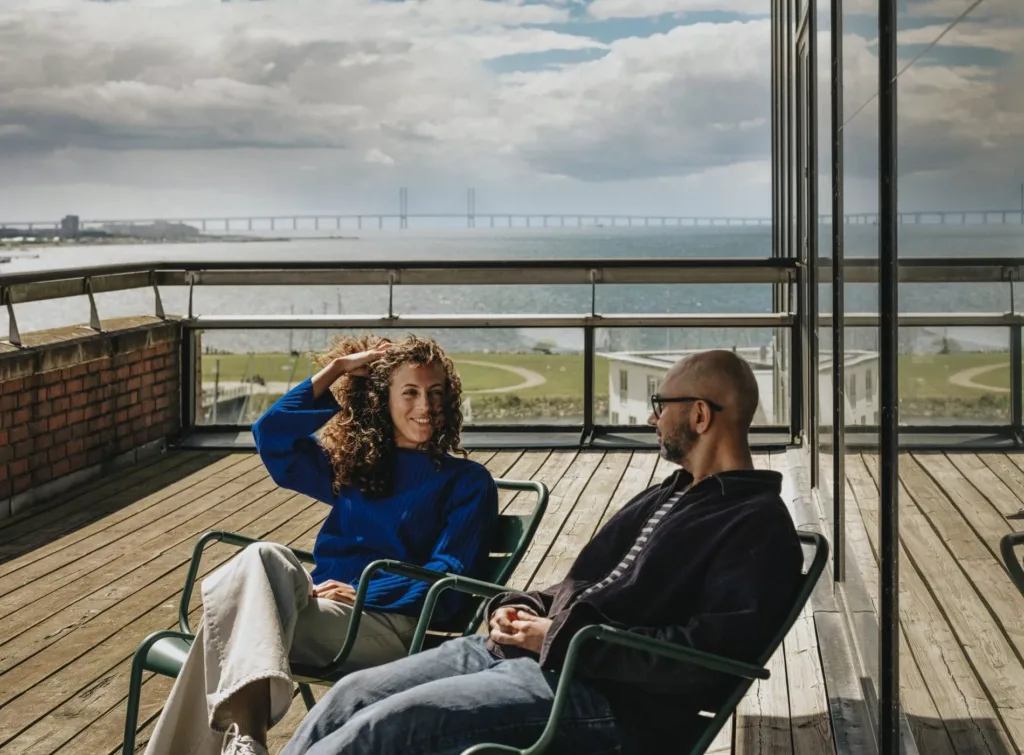 Två personer sitter på en terrass med utsikt över en bro och havet.