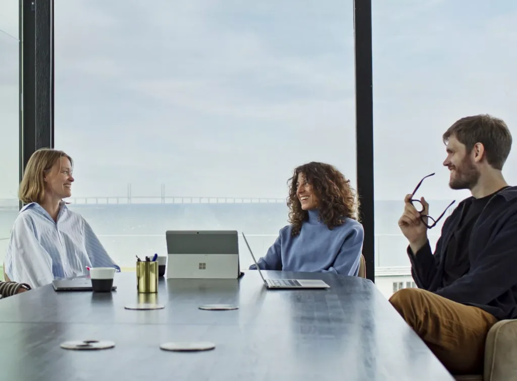 Tre personer sitter vid ett konferensbord med utsikt över havet i bakgrunden, och en av dem håller i sina glasögon.