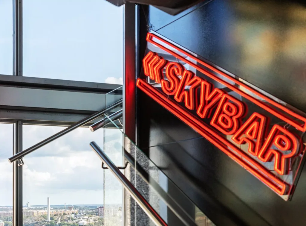 En röd neonskylt med texten "Skybar" vid en glasvägg med utsikt över en stad.