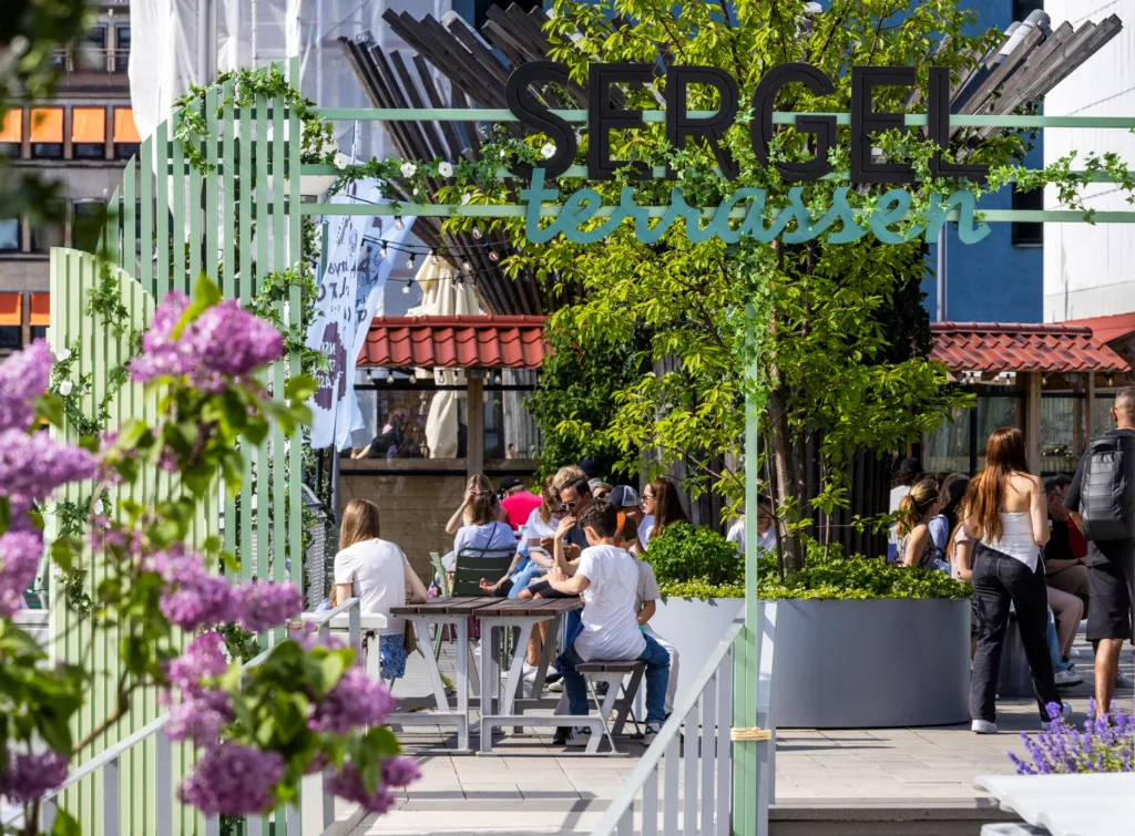 3. Entré till "Sergel Terrassen" med grönskande växtlighet, lila blommor och människor som sitter vid bord i bakgrunden.