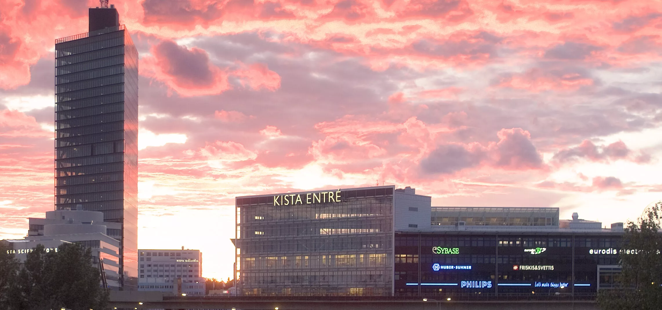 Kista Entré-byggnaden mot en rosa och orange solnedgång med företagsskyltar på fasaden.