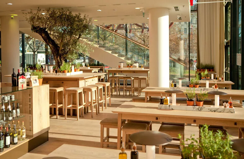 En ljus och modern restaurang med träbord, stolar och gröna växter i en rymlig miljö.