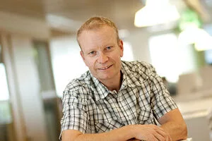 En man med kort blont hår och rutig skjorta lutar sig mot en bordsskiva och ler.