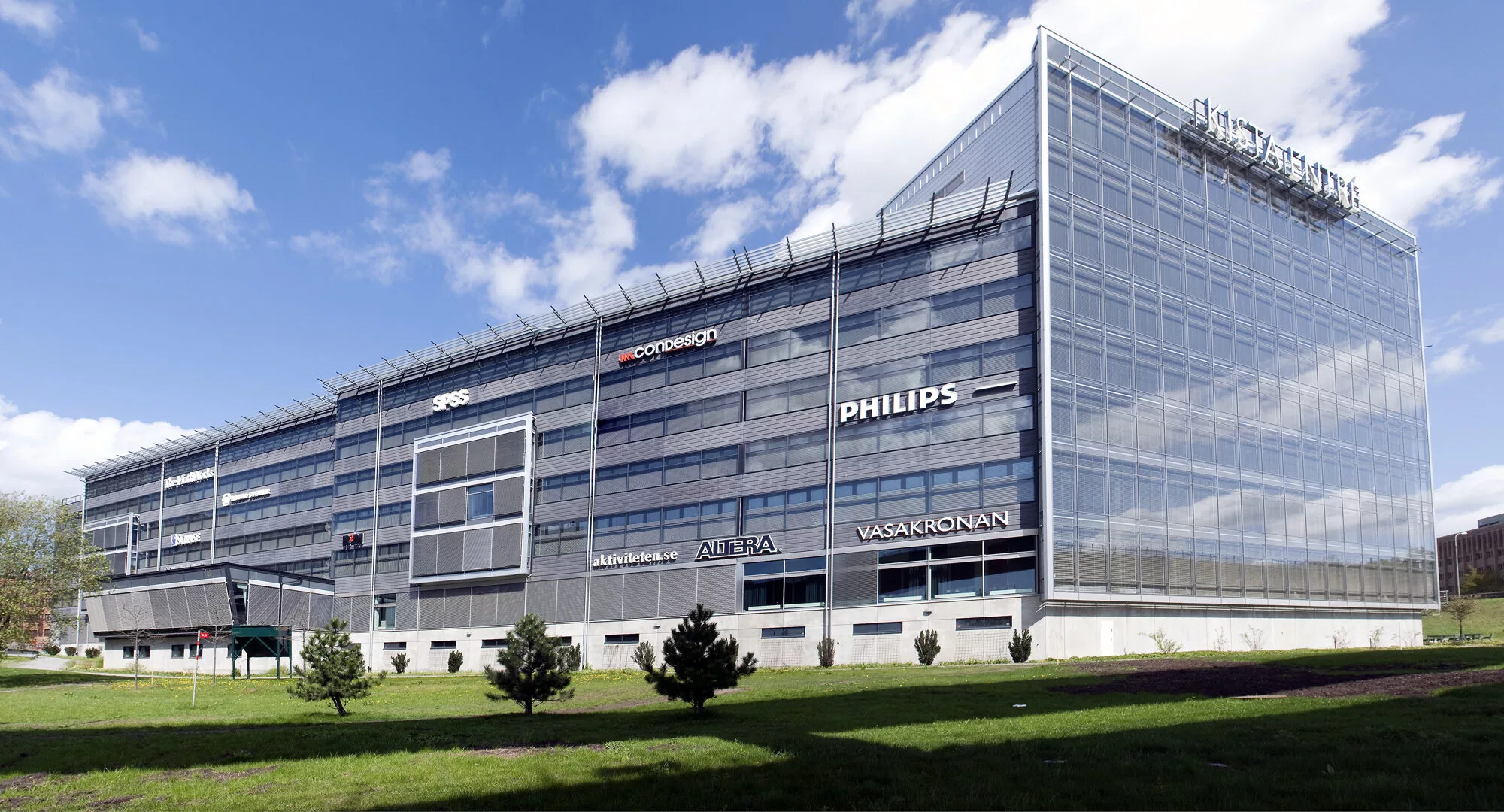 1. En modern byggnad med glasfasad, tydliga företagslogotyper som "Philips" på utsidan, omgiven av grönska och träd under en blå himmel med moln.
