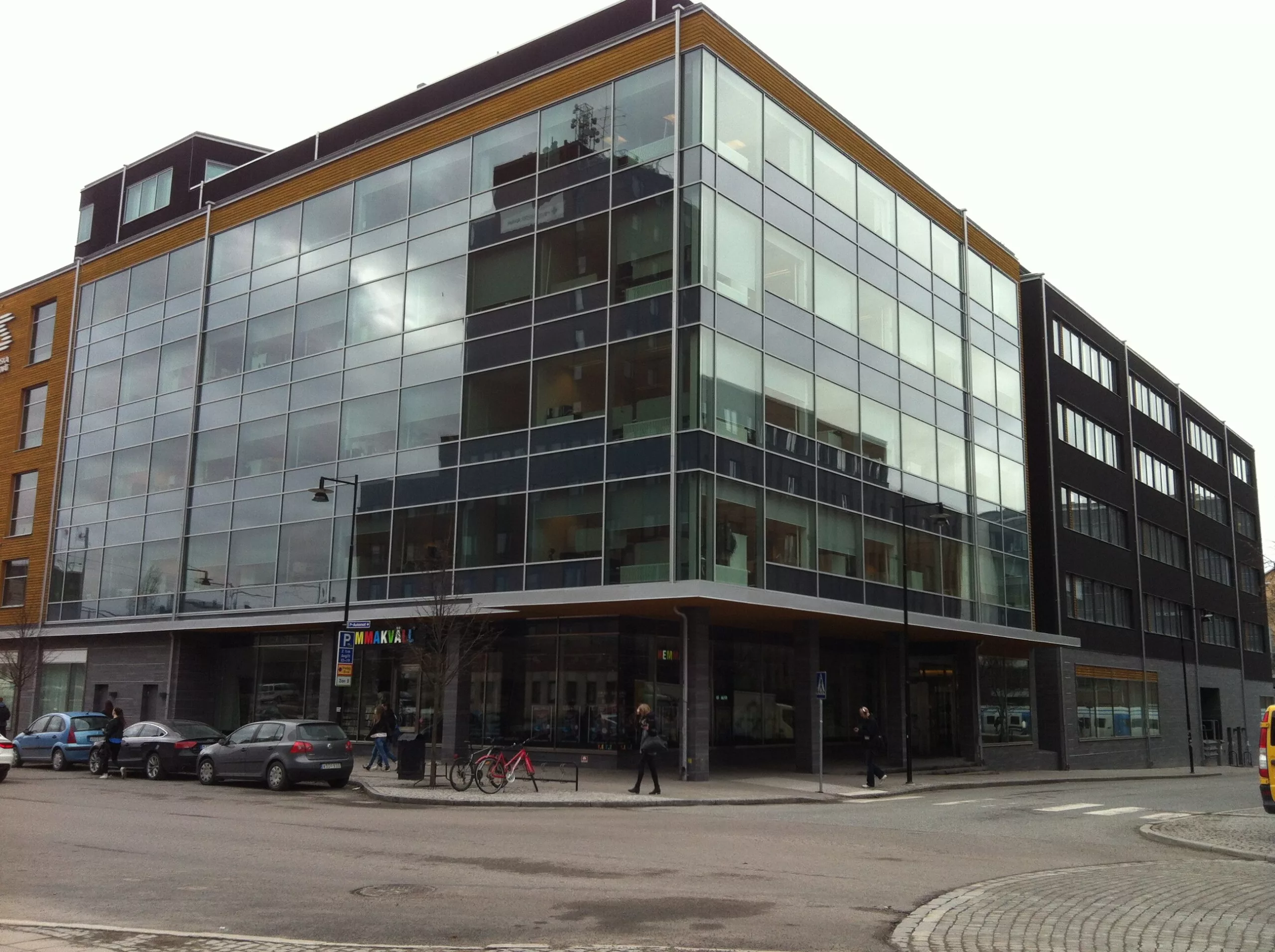 4. En modern glasbyggnad vid en gata, med parkerade bilar och trottoarer runt omkring.