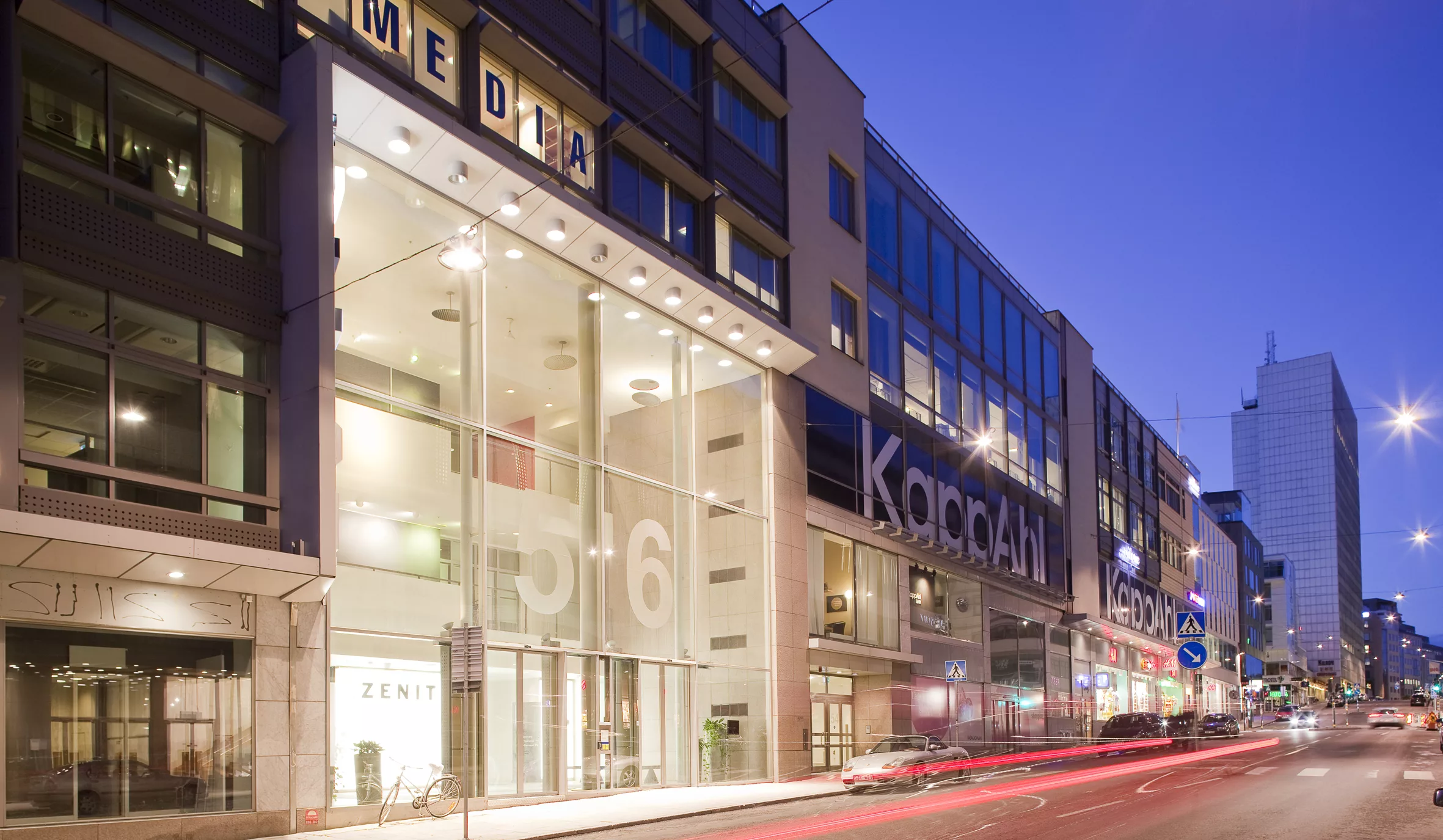 En modern byggnad med glasfasad och skyltar, föreställande "Kongahälla Center", belyst under kvällstid.