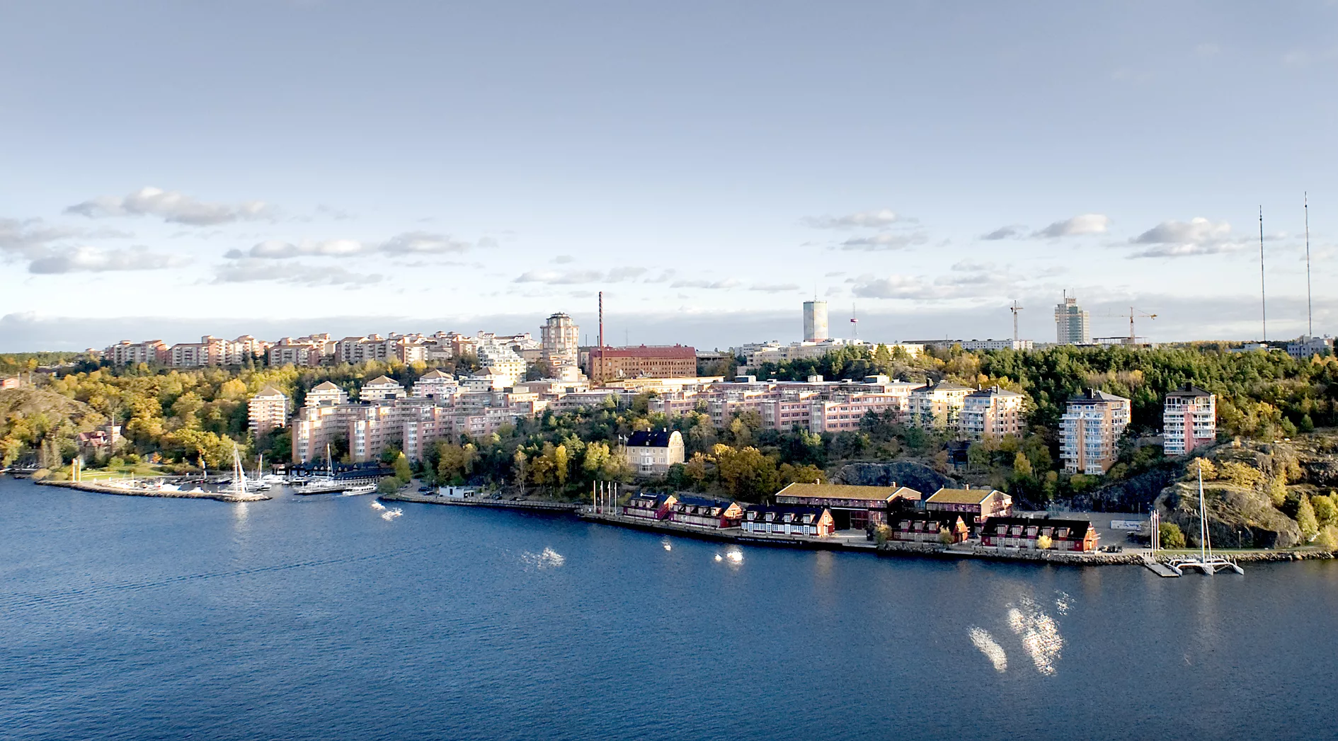 En stadsvy med vatten i förgrunden, byggnader omgivna av grönska och en klar himmel.