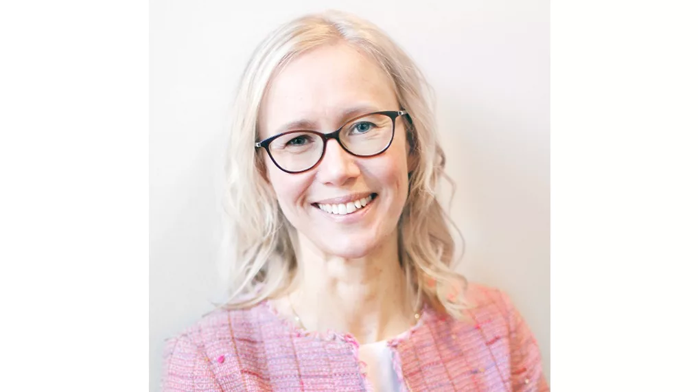 En kvinna med blont hår, glasögon och rosa kavaj ler mot en ljus bakgrund.