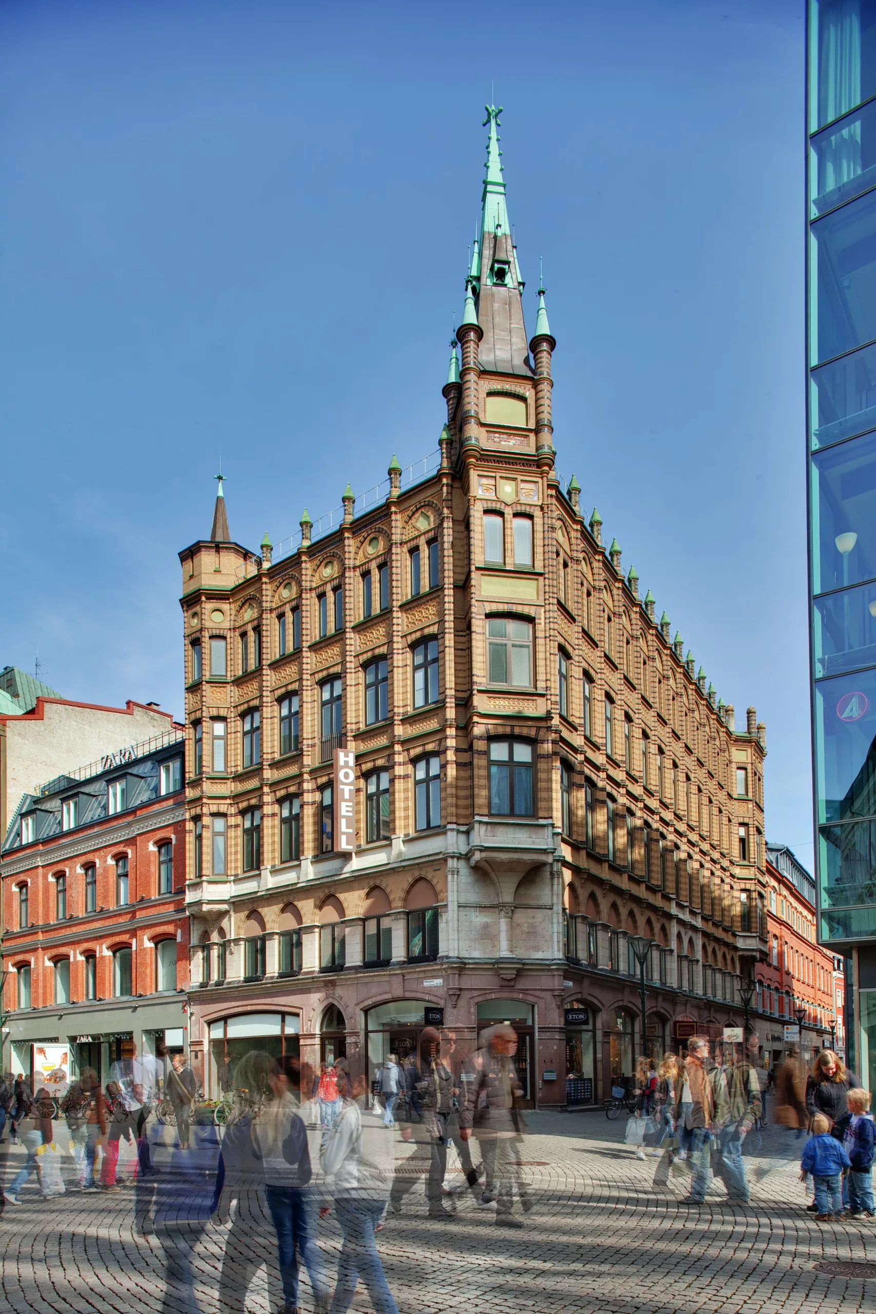 1. En historisk byggnad med tegelarkitektur och spetsiga torn, omgiven av människor och en modern glasbyggnad i bakgrunden.