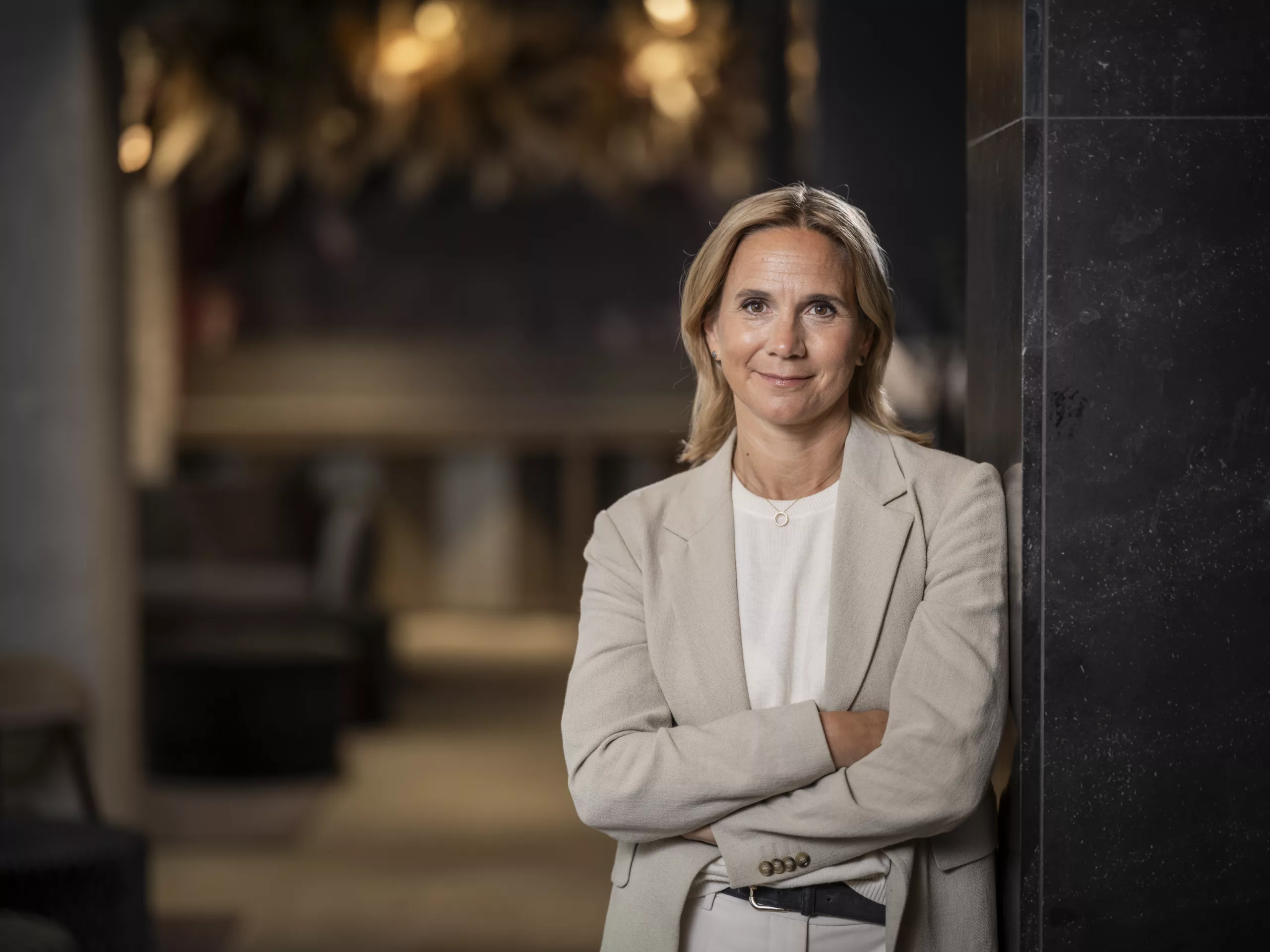En kvinna i ljus kavaj står med armarna i kors i en elegant inomhusmiljö.
