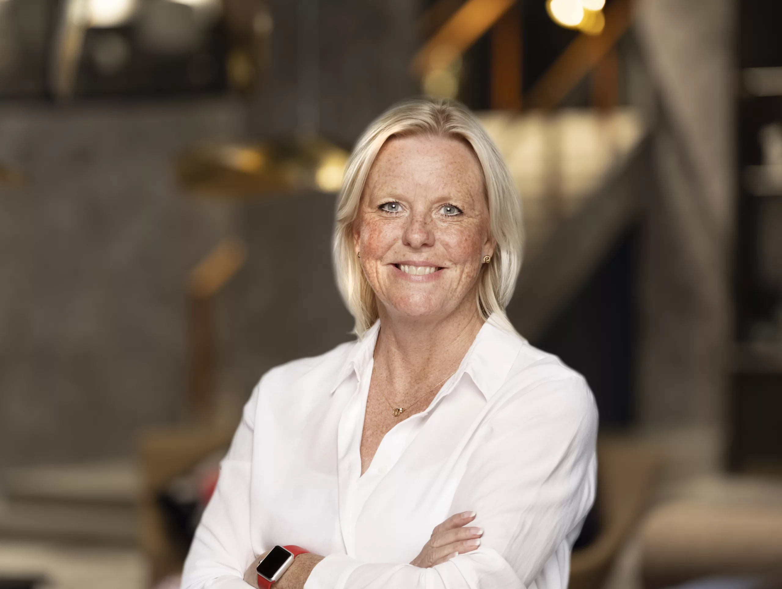 En kvinna med blont hår och vit skjorta ler och står med armarna i kors framför en suddig bakgrund.